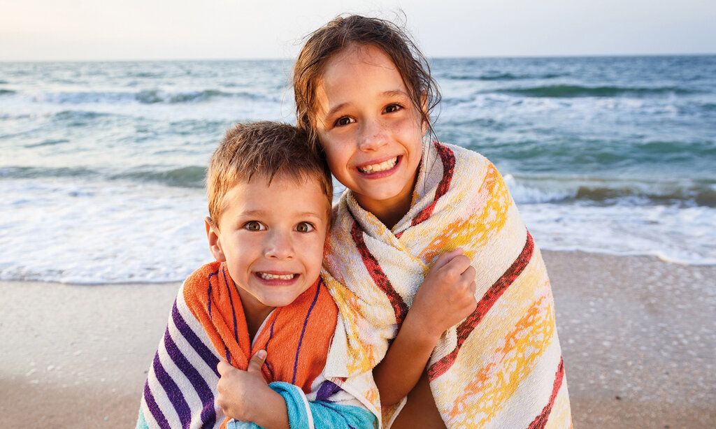 Ein Junge und ein Mädchen am Strand, in Handtücher gehüllt, grinsen in die Kamera, im Hintergrund Meer