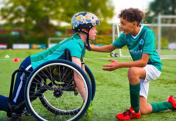 Filmszene aus Bleib am Ball, ein Junge mit Helm kniet in seinem Rollstuhl, ein andere kniet vor ihm und redet auf ihn ein, beide sind auf einem Fußballplatz