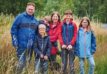 Porträt von familie Teichmann aus Grevenbroich. Vater, Mutter, drei Jungs, ein Mädchen und ein Hund stehen auf einer hochgewachsenen Wiese. Die Mutter trägt den kleinsten Sohn auf dem Arm, der weint.