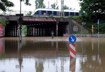 Hochwasser auf einer Straße in Düsseldorf, Straßenschilder stehen im wasser, im Hintergrund eine Unterführung, darauf eine S-Bahn