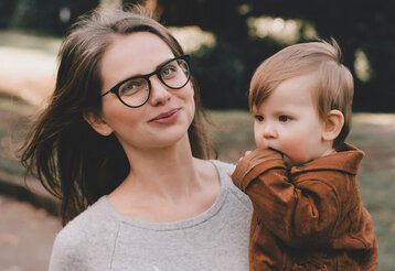 Porträt einer jungen Frau mit Brille, die ihr Kleinkind auf dem Arm trägt, im Hintergrund herbstlicher Park