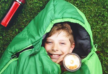 Blick von oben auf einen Junge, der mit Taschenlampe im Schlafsack liegt, Untergrund: Wiese