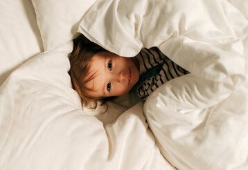 Kind liegt im Bett und guckt unter der Bettdecke hervor