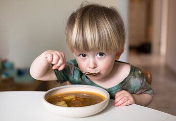 Kleines Kind sitzt an einem Tisch und isst Suppe, guckt dabei in die Kamera