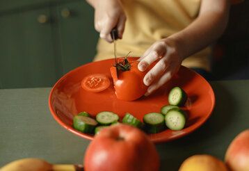 Detailaufnahme, ein Kind schneidet auf einem roten Teller eine Tomate, geschnittene Gurken liegen auf dem Teller, auf der Arbeitsplatte liegen Äpfel und Bananen