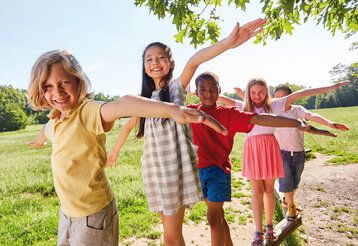 Fünf Kinder balancieren auf einem Holzbalken, im Hintergrund eine Wiese und Bäume