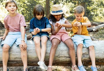 Vier Kinder sitzen auf Baumstamm und gucken auf ihre Armbanduhren