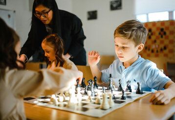 zwei Kinder spielen Schach, im Hintergrund eine Frau, die mit einem Mädchen spricht