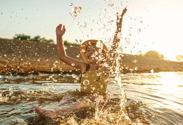 ein Mädchen mit einem Sonnenhut planscht im See, Wasser spritzt