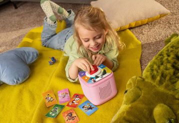 Mädchen liegt mit Toniesbox und Spielkarten auf einer Decke auf dem Boden