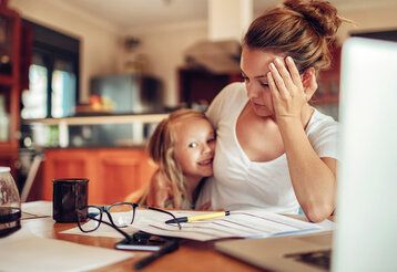 Mutter sitzt am Schreibtisch und ist genervt, kleine Tochter im Arm, die lacht