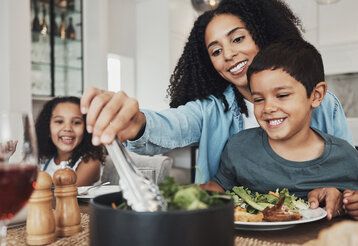 Eine Mutter sitzt mit Tochter und Sohn am Esstisch und tut Salat auf einen Teller