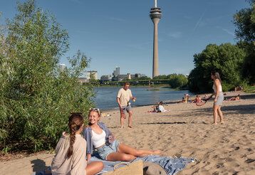 Paradiesstrand Düsseldorf, zwei Frauen sitzen im Sand, ein Man und eine Frau spielen Beachball, im Hintergrund der Fernsehturm