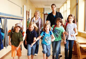 fünf Kinder rennen durch den Flur einer Schule, der Kamera entgegen, hinter ihnen gehen eine Lehrerin und ein Lehrer