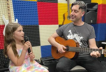 Ein Mädchen sitzt mit seinem Musiklehrer zusammen. Das Mädchen singt in ein Mikrofon, der Lehrer spielt Gitarre. Im Hintergrund sieht man, dass der Raum schallgedämpft ist, die Wände sind mit blauen, roten, schwarzen ud weißen Platten verkleidet, Musikinstrumente, Verstärker und Boxen sind zu sehen