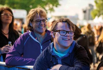 Junge mit Down-Syndrom steht lachend im Publikum, hinter ihm zwei Frauen