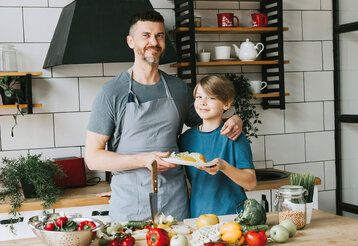 Vater und Sohn stehen in der Küche, vor ihnen liegt Gemüse, sie halten zusammen einen Teller, der Vater hat den Arm um den Sohn gelegt