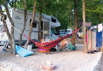 Ein Wohnmobil steht auf einem Campingplatz, zwischen Bäumen ist eine Hängematte gespannt, Familie sitzt vor Wohnmobil