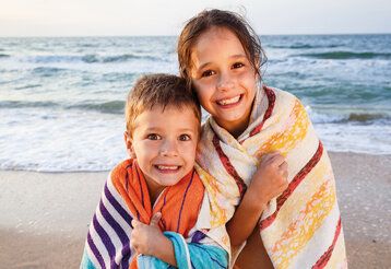 Ein Junge und ein Mädchen am Strand, in Handtücher gehüllt, grinsen in die Kamera, im Hintergrund Meer