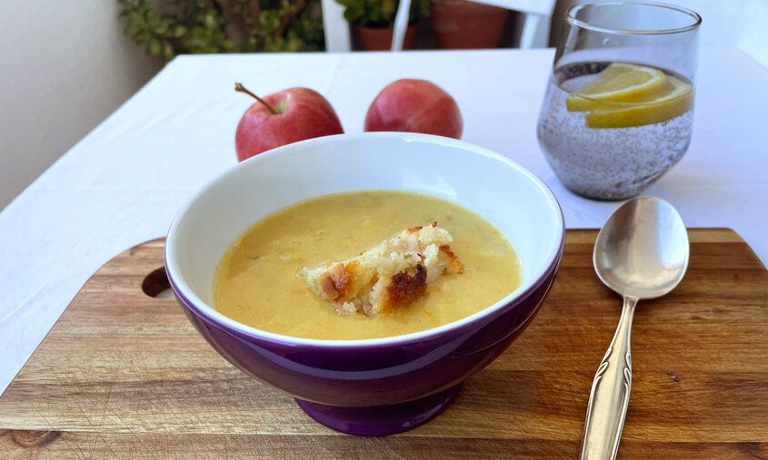 Eine Suppenschale mit Kartoffelsuppe und einem Stück Apfelkuchen steht auf einem Tisch mit einer weißen Tischdecke, daneben liegt ein Löffel und ein Wasserglas steht auch auf dem Tisch